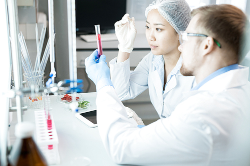 Crop side view of young scientists sitting at laboratory desk and analyzing nutrition properties of solution in test tube