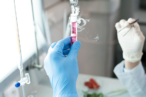 Crop hands of microbiologists studying test tube with pink emitting smoke solution in it