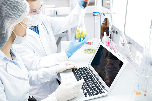 High angle portrait of two modern scientists working on food research studying liquids in beaker while sitting at table in laboratory and using laptop