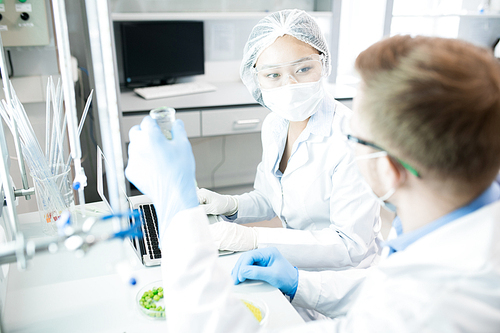 Microbiologists in protective laboratory clothes sitting at table and looking at each other discussing properties of green vegetables being tested, crop man showing woman flask with samples