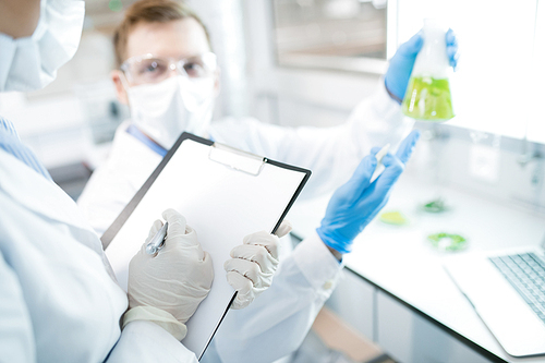 Crop female microbiologist writing down into clipboard test results of research of male holding flask with green solution and pointing at it sitting at desk