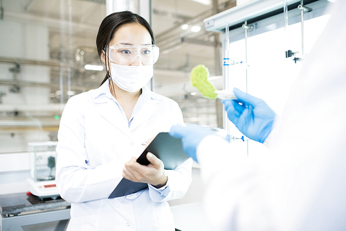 Crop back view of microbiologist holding in hands pincers with green food nutrition test sample and woman looking at it writing down results into clipboard