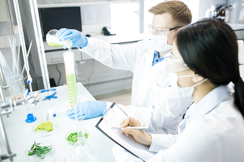 Scientists pouring green solution from flask into measuring cylinder and writing down results of chemical reaction with green vegetable samples in bowls