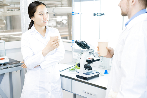 Crop view of smiling microbiologist man and woman standing together at desk with microscope and drinking hot beverage smiling and discussing results of recent testing