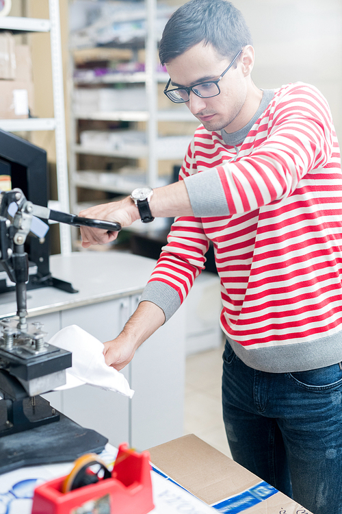 Casual man working in modern printing office and using press to create print on white cap