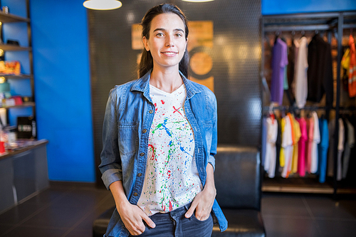 Happy attractive saleswoman in denim shirt and fashionable tshirt with paint splashes holding hands in jeans pockets and , she working in youth store