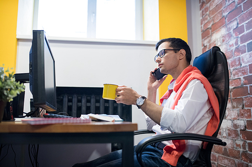 Serious pensive young male manager in glasses sitting at desk and drinking coffee while talking by phone in loft office