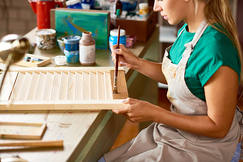 Side view of blonde young woman enjoying work in art studio painting shutters with bronze paint, making DIY interior decorations