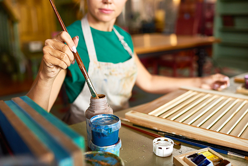 Mid-section portrait of pretty blonde woman enjoying work in art studio painting picture frame with paint, focus on female hand dipping brush in paint