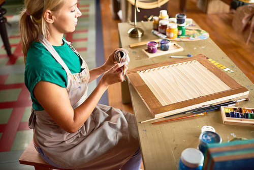 Side view portrait of young blonde woman enjoying work in art studio painting shutters with bronze paint, making DIY interior decoration