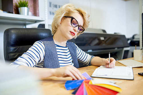 Attractive young woman in glasses looking at color palette and writing in notebook while sitting at table in office.
