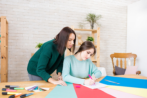 Creative interior designer wearing turquoise sweater sitting at wooden desk and presenting ideas with help of marker pen and sheet of paper while her female colleague keeping eye on her.