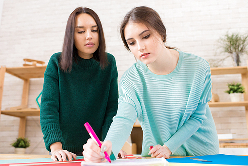 Attractive young student wearing knitted sweater using felt-tip pen and sheet of paper in order to share ideas with female groupmate while working together on university project.