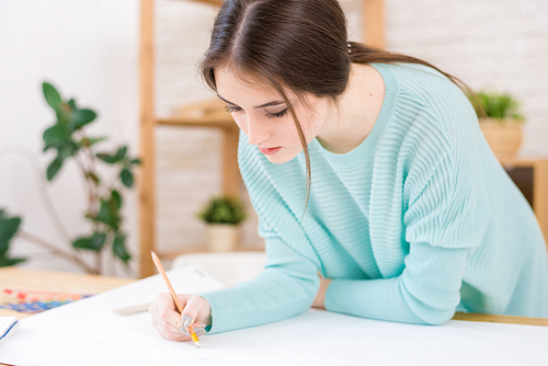 Concentrated young architect wearing turquoise sweater working on blueprint while leaning on wooden desk, office interior on background