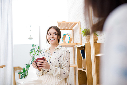 Coffee break in modern design studio: pretty young woman sitting with cup in hands and chatting animatedly with her colleague