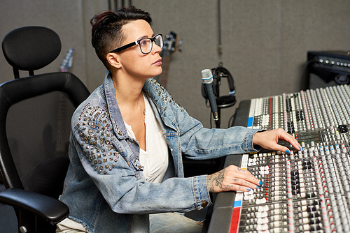 Side view of stylish woman in denim using control board while working in sound studio.