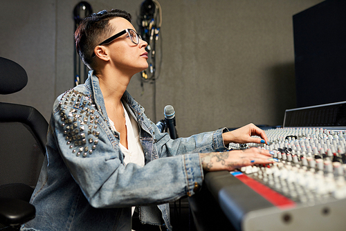 Concentrated confident hipster female sound designer in eyeglasses using mixing desk while working in modern recording studio