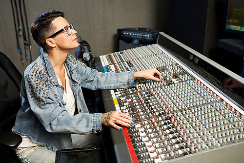 Side view of tattooed stylish woman in denim jacket using music control board while working in studio.