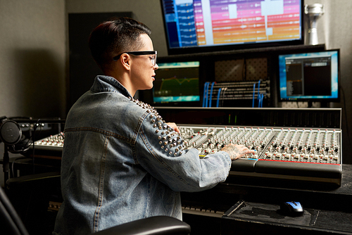 Pensive busy hipster young woman in denim jacket working on professional analog mixing console while creating unique soundtrack in recording studio