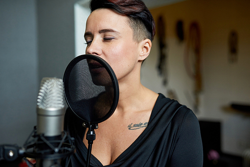Beautiful sensual woman singing in music recording studio standing near microphone with eyes closed.