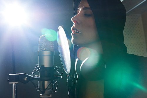 Young sensual woman recording song in studio while singing with eyes closed into mic.