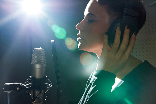 Side view of beautiful girl with eyes closed wearing earphones while singing song with microphone in studio.