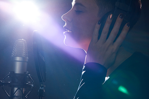 Sensual girl wearing headphones and singing with microphone in bright light keeping eyes closed.