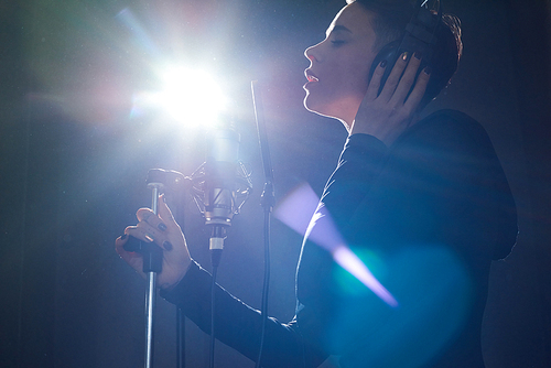 From below side view of alluring girl wearing headphones while singing into microphone in bright light of recording studio.