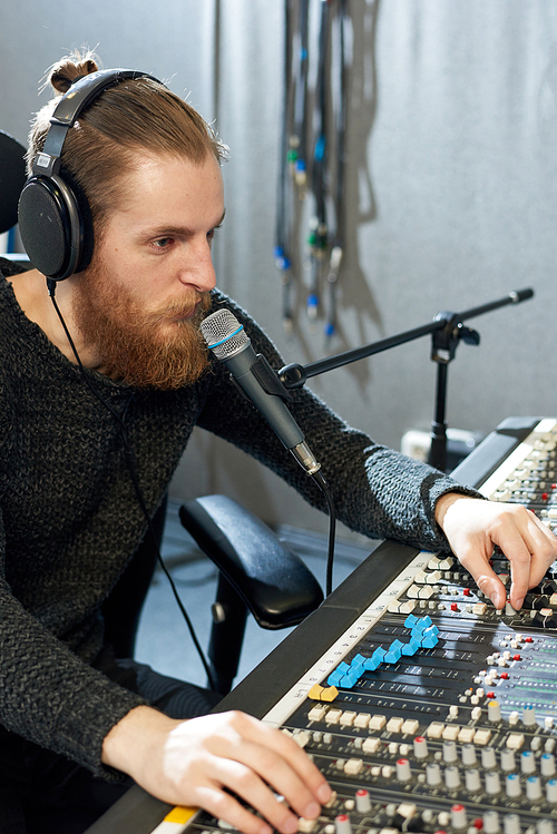 Casual bearded man in headphones speaking into microphone while using music console in sound studio.