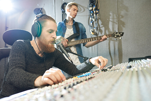 Serious handsome hipster male audio engineer with beard working on digital audio workstation while listening to music in headphones, young guitarist playing musical instrument while recording song