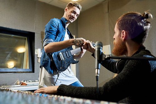 Young trendy man with guitar and studio worker hitting with fists looking excited with great job done in recording studio.