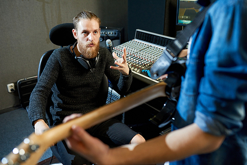 Bearded man sitting at console in recording studio and talking to crop man with guitar preparing new song.