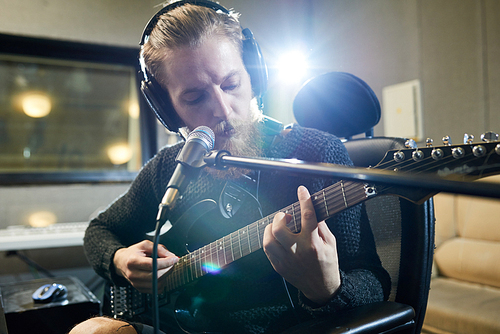 Serious creative hipster musician in headphones playing guitar and singing song while recording new hit in studio, man in sunlight