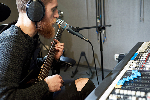 Side view of man in headphones playing guitar and singing into microphone sitting at console in recording studio.