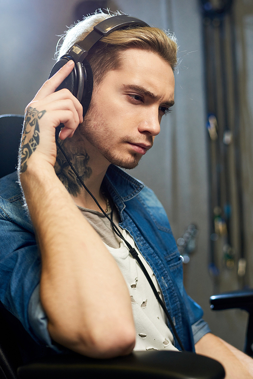Tattooed young man in earphones listening to new composition in recording studio looking serious.