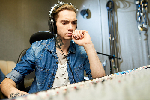 Young trendy man wearing headphones and listening to recording of new song sitting in chair in sound studio.