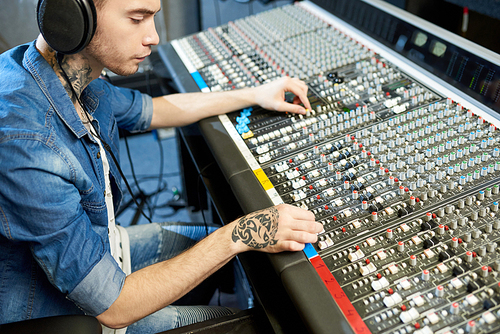Side view of man with tattoos wearing headphones and using console to edit music recording working in studio.