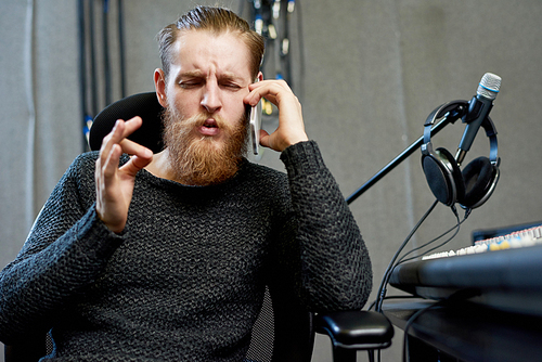 Bearded man sitting in armchair inside of music recording studio and having phone call.