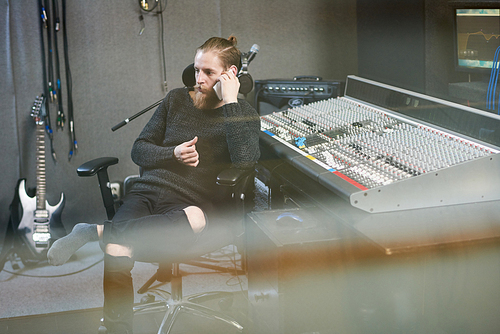 Bearded stylish operator sitting in modern recording studio and having call on smartphone.
