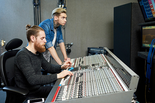 Side view of trendy men sitting at huge console and working on new musical project in recording studio.