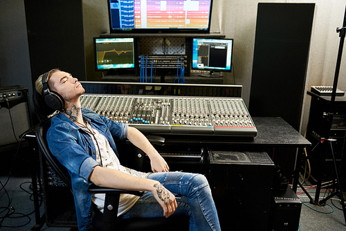 Side view of stylish man in headphones listening to music with enjoyment sitting at console in recording studio.
