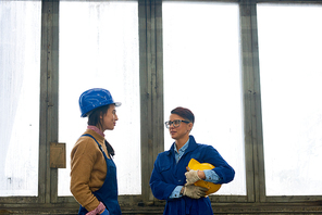 Waist up portrait of two female workers chatting standing against window in industrial workshop, copy space