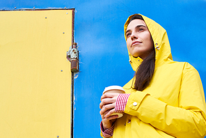 Bright colorful portrait of young woman wearing yellow raincoat standing against contrasted blue wall holding coffee cup and looking away, copy space