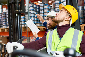 Side view portrait of young man sitting in forklift car looking up and moving goods with supervisor giving instructions in background, copy space