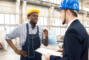 Bearded African American factory worker wearing hardhat and overall discussing results of accomplished work with his superior while standing at production department