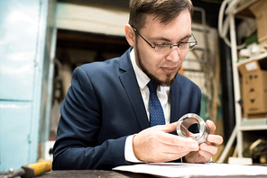 Waist-up portrait of handsome young inspector wearing eyeglasses and suit studying steel workpiece with concentration while carrying out quality control at production department of modern plant.