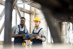 Multi-ethnic group of machine operators wearing overalls looking away pensively while taking coffee break in spacious production department of modern plant, one of them holding digital tablet