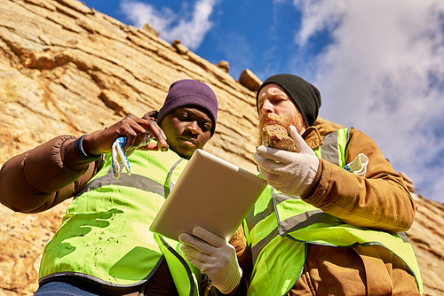 Low angle portrait of two tough workers wearing reflective jackets, one of them African, inspecting mineral ore on site outdoors and using digital tablet