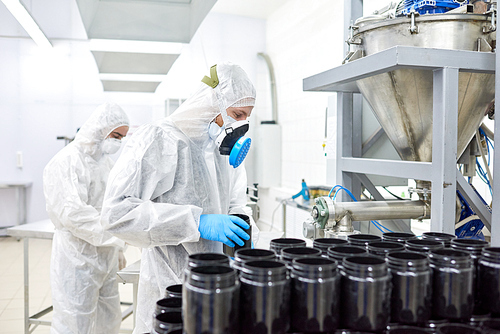 Group of concentrated factory workers wearing coveralls, respirators and rubber gloves filling in plastic bottles with sport supplement
