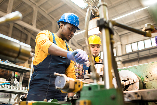 Multi-ethnic team of workers wearing overalls and protective helmets using lathe in order to machine workpiece, interior of spacious production department on background